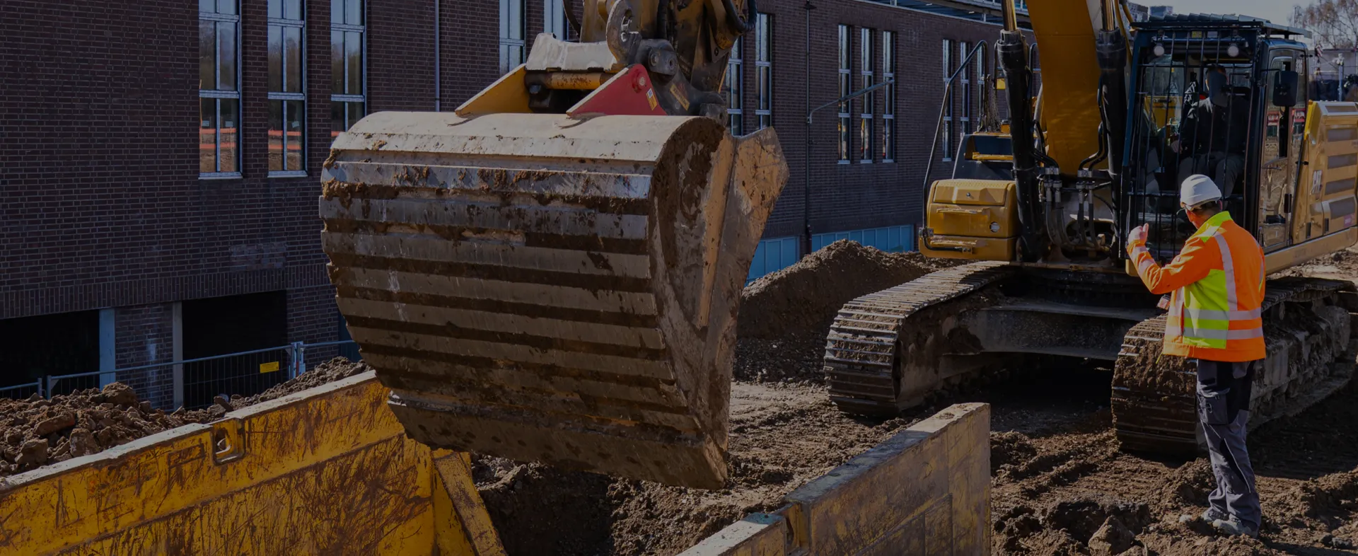 Bagger mit großer Schaufel auf einer Baustelle vor einem Backsteingebäude; daneben steht eine Person in orangefarbener Warnschutzkleidung und weißem Schutzhelm.