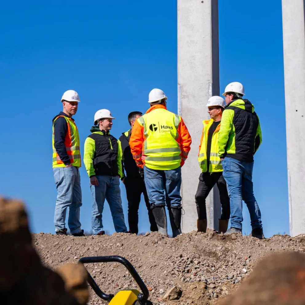 Mehrere Bauarbeiter in Warnschutzkleidung und Schutzhelmen stehen auf einer Baustelle vor hohen Betonstützen und klarem blauem Himmel.