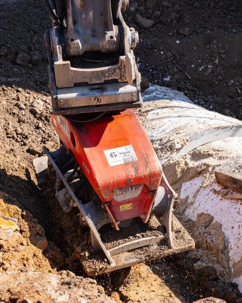 Rotes Verdichtungsgerät mit Metallplatte in einer ausgehobenen Baugrube auf einer Tiefbaustelle