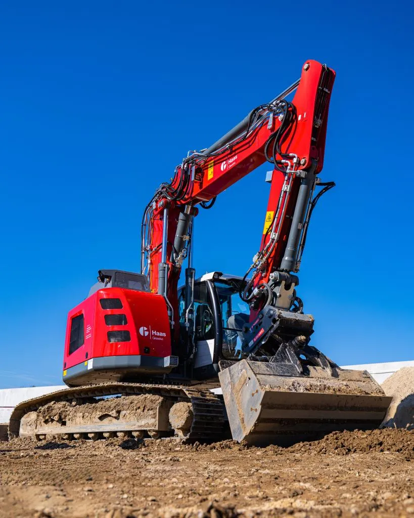 Roter Bagger mit großer Schaufel auf einer Baustelle vor strahlend blauem Himmel.