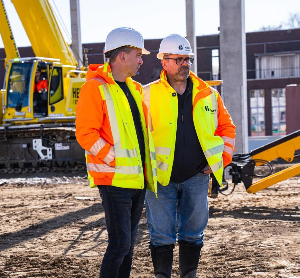 Zwei Bauleiter in orange-gelben Warnschutzjacken und weißen Schutzhelmen stehen auf einer Baustelle vor einem gelben Bagger
