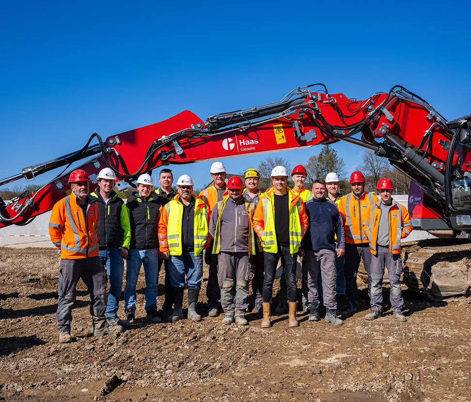 Gruppe von Bauarbeitern in Warnschutzkleidung vor einem großen roten Haas-Gieseke-Kettenbagger auf einer Baustelle